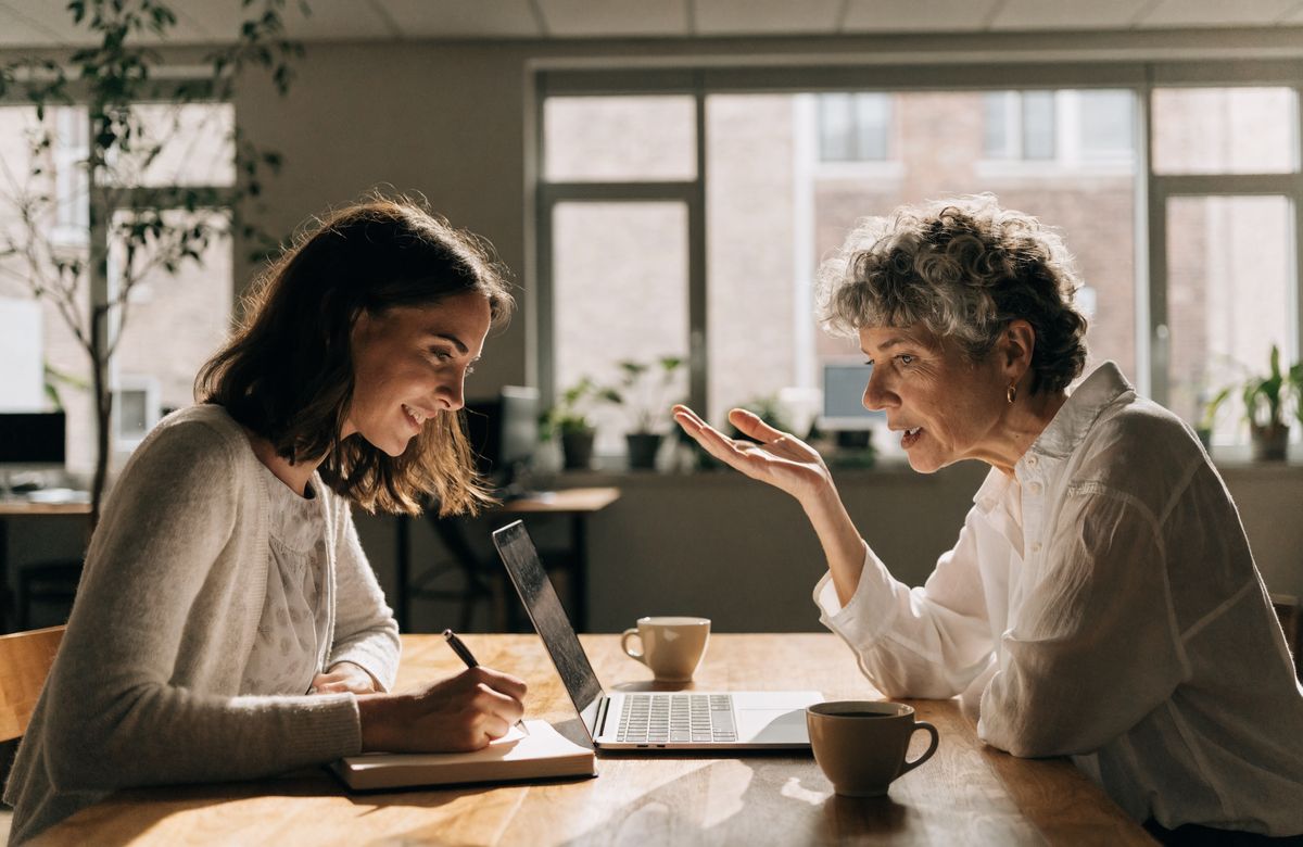 Zwei Frauen im Gespräch am Tisch: Eine erklärt, die andere macht sich Notizen.